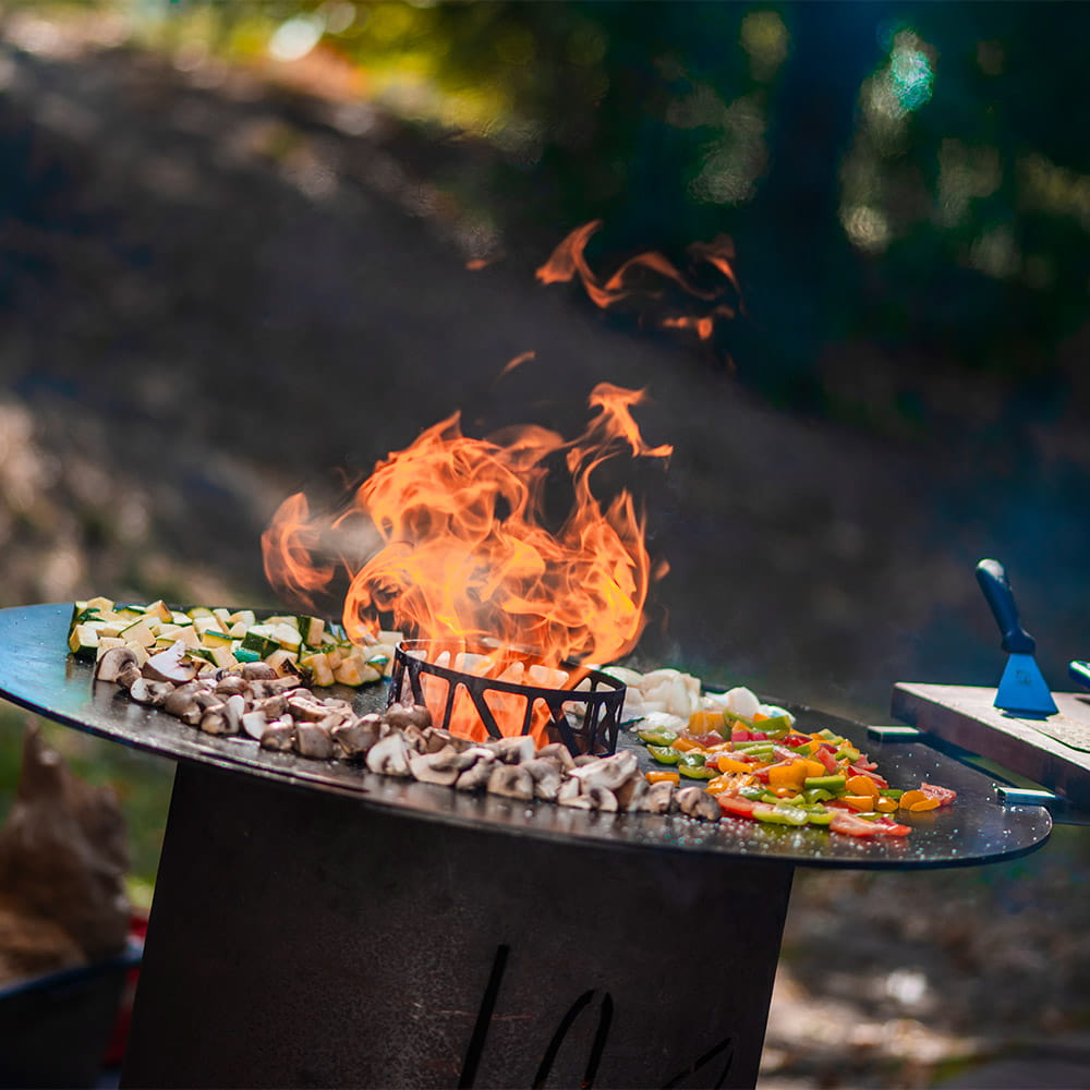 Feuerplatte im Freien mit Champignons, Zucchini und Paprika, in der Mitte lodert das Feuer